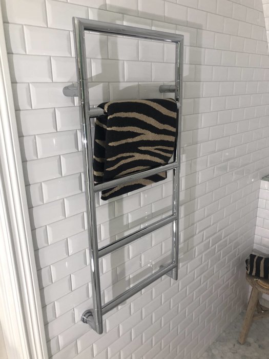Nyinstallerad kromad handdukstork på vit kaklad badrumsvägg med en svart och beige randig handduk.