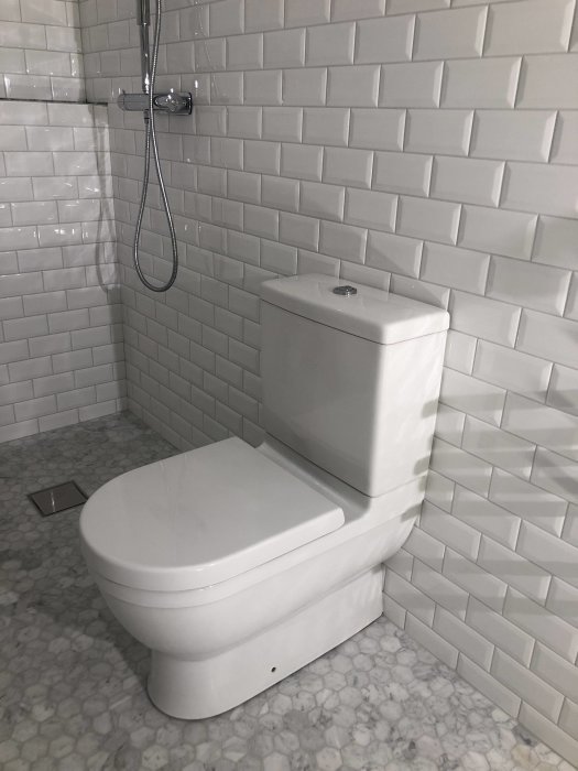 Nyinstallerad vit toalett intill en dusch med vita metrokakelväggar och marmormönstrat golv.