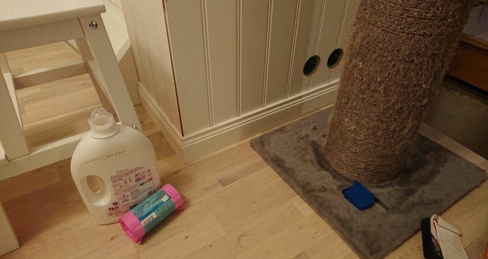 Ett hörn av ett rum med trägolv, en kattklöspelare, tvättmedel och rengöringsprodukter på golvet.
