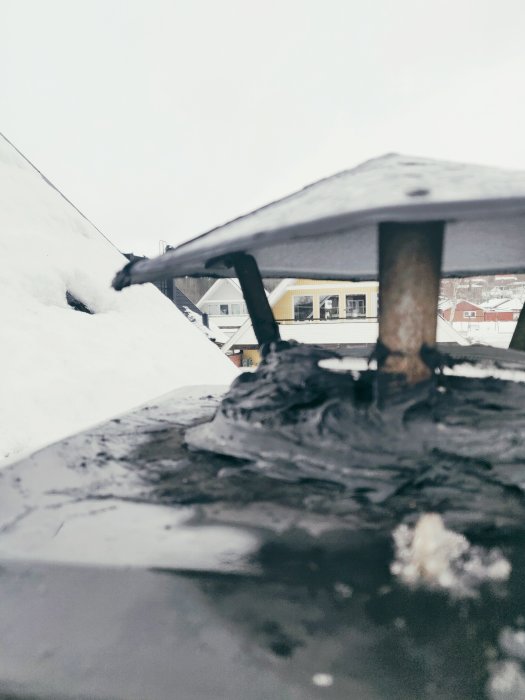 En takhuvsöverdel täckt med tjockt lager av svart tätningsmedel på ett snötäckt tak, med oskarp bakgrundsbild av hus.