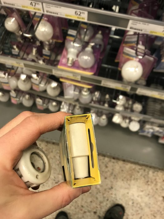 En hand håller en gammal säkring framför säkringsutbudet i en affär.