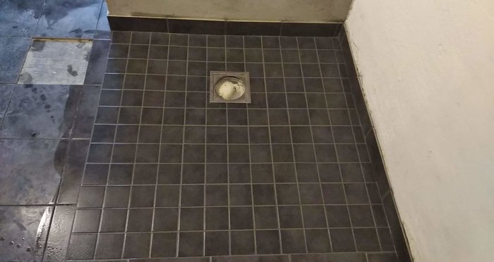 Nyfogade mörkgrå klinkergolv med synlig golvbrunn, nära en vit vägg och blå skyddsplast.