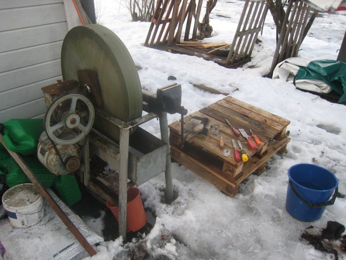 En grov slipsten monterad utomhus täckt av snö, med olika verktyg på en träpall framför.