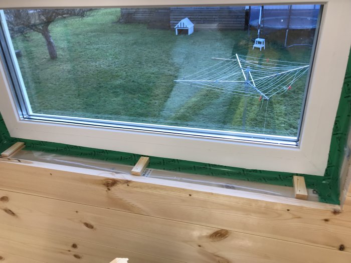 Träpanel nära fönster under installation, synlig tilluftsslang och grön tätningsduk, utsikt mot trädgård.