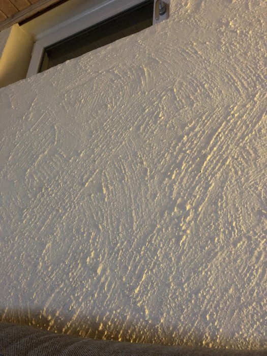 Texturerad kalkborstad vägg med synliga borstslag nära ett fönster, vissa delar med flagnande färg.