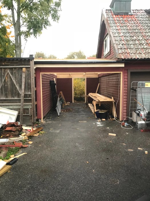 Carport under konstruktion med takstöd och rödmålade väggar, utan grindar och dörr, byggbråte i förgrunden.