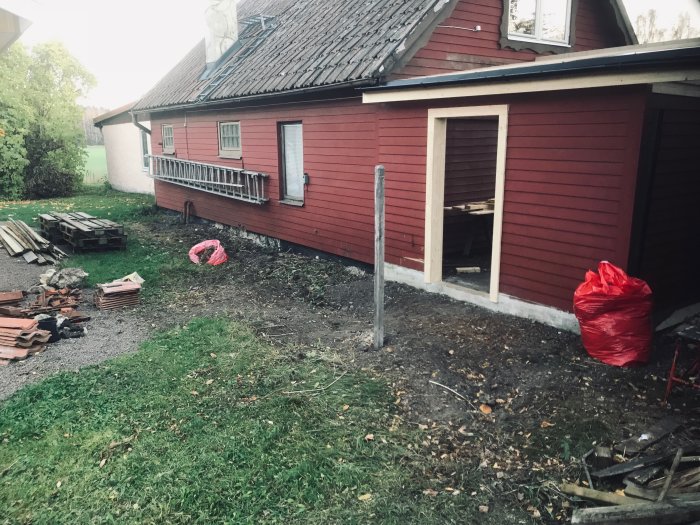 Rödfärgad byggnad sett från baksidan med pågående arbete på ett nytt carporttak, omgiven av byggmaterial.