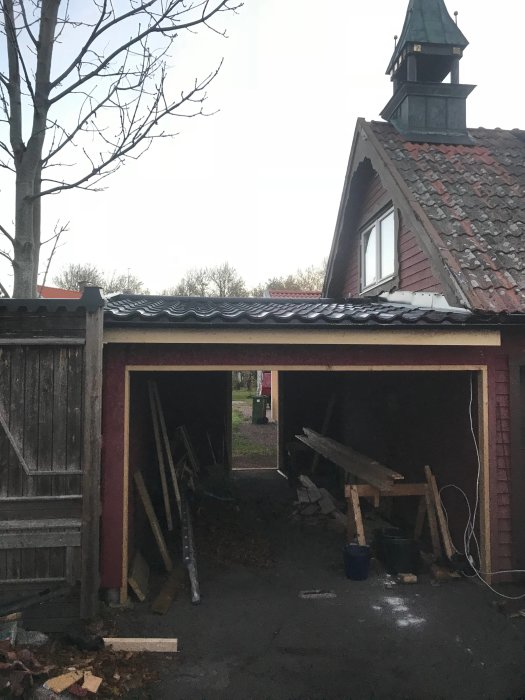 Halvfärdig carport från baksidan med tak, utan grindar och dörr, byggmaterial synligt, nära ett rött hus.