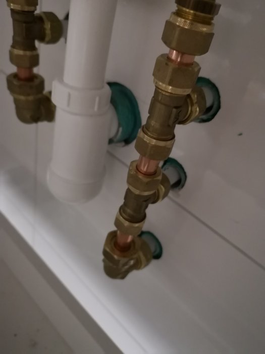 Vattenrör i koppar och mässing utan silikonfog där de går in i vit vägg bakom kommod.