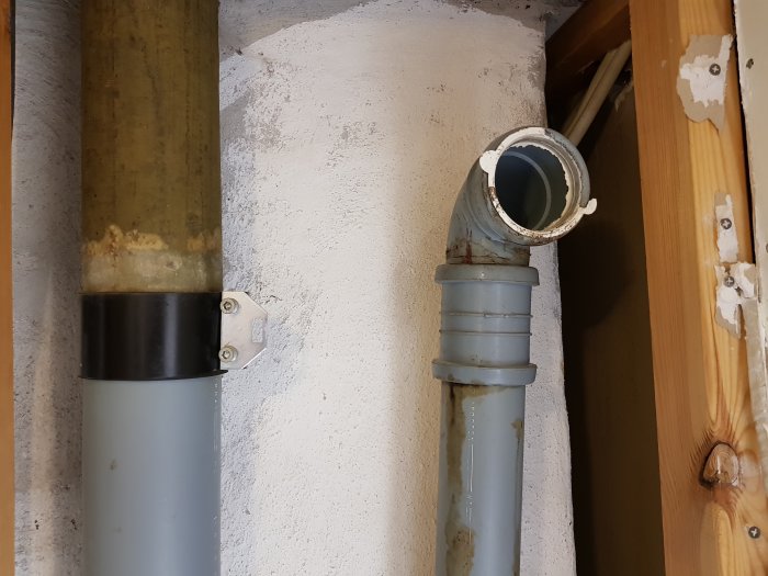 En öppen avloppsrörskoppling i en vägg, en pluggad avloppsstam intill träreglar, tecken på skada.
