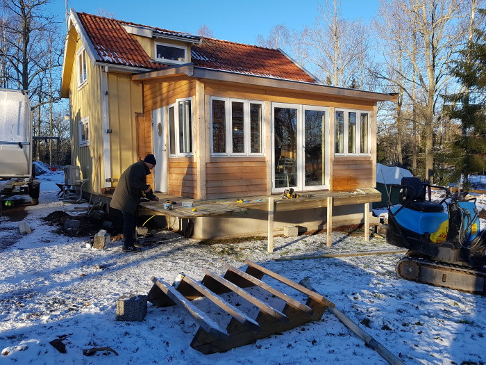 Pågående arbete på trädäck vid en vitoljad glasveranda på en solig dag med snö på marken.