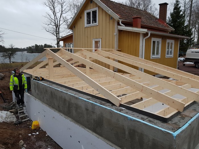 Delvis färdigställd isolergrund med påbörjad återfyllning och träkonstruktion för trädäck vid ett gult hus.