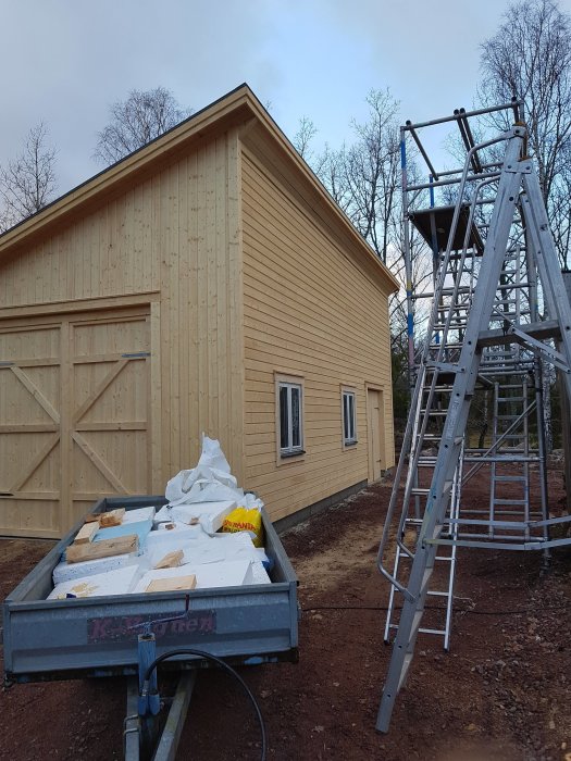 Isolergrund och nybyggt garage med fönster, träpanel på glasverandan, samt byggmaterial och stege.