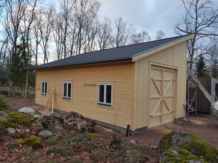 Nybyggt garage med fönster och träpanel nära en glasveranda mot en skogsbakgrund.