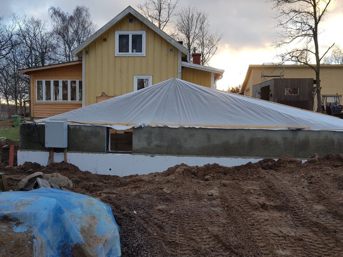 Isolergrund till ett hus med dränering i fokus och återfyllning pågår, gult hus med glasveranda i bakgrunden.