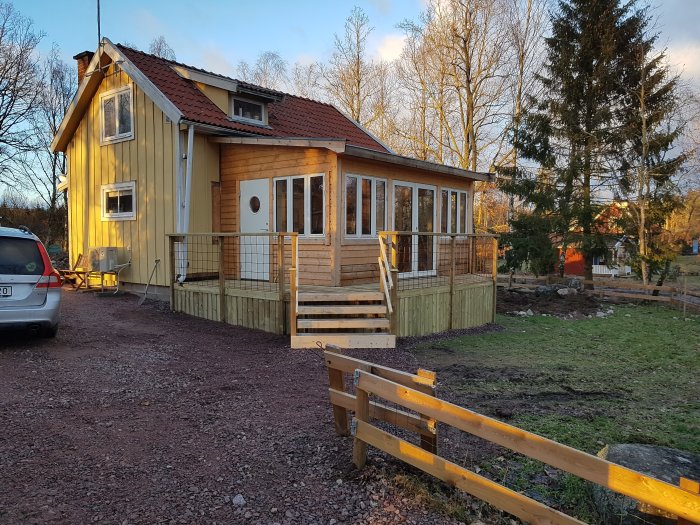 Ett nyrenoverat hus med gult träslag, glasveranda med träpanel, och nybyggt trädäck i solnedgångsljus.