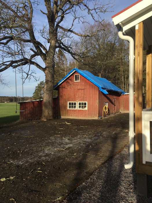 Gammal röd verkstad med oisolerat 1 1/2 plan under en blå presenning på taket, belägen på en lantlig tomt.