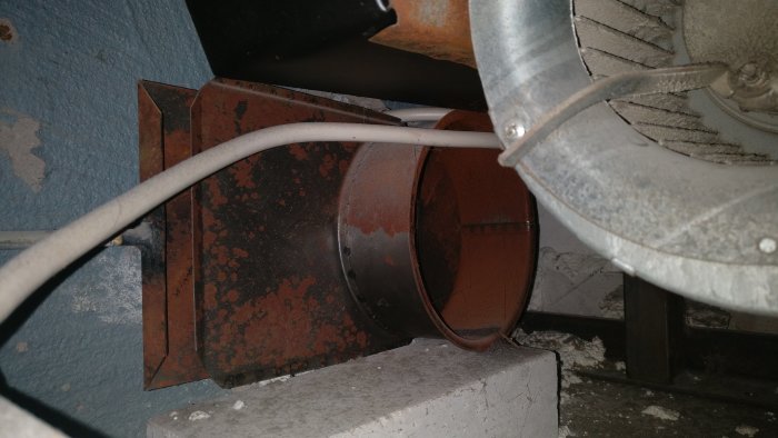 Rostigt spirorör i en källare kopplat till en braskaminsfläkt, med synliga fläktdelar och kablar.