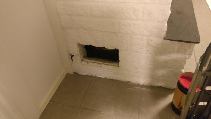 Fläktens luftintag i källare med svart spjäll synligt genom rektangulärt hål i vitmålad vägg.