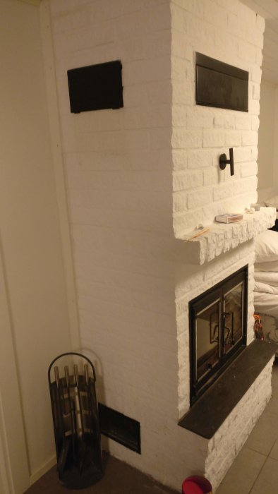 Vitmålad braskamin med luftfläkt och spjäll i källare, inklusive verktygsställ och luftintag.