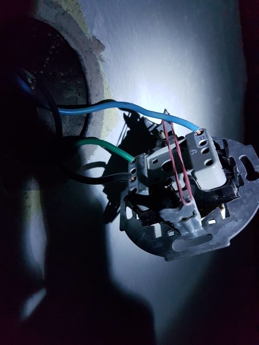 Närbild av en elektrisk kopplingsdosa med anslutna kablar i svart, blått och gröngult över en väggbrytare.