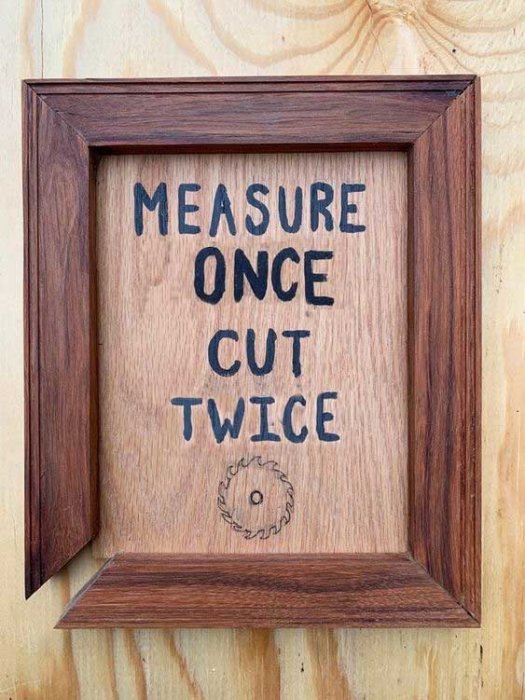 Träram med texten "MEASURE ONCE CUT TWICE" och ritning av sågblad, humoristisk omtolkning av snickarordspråk.