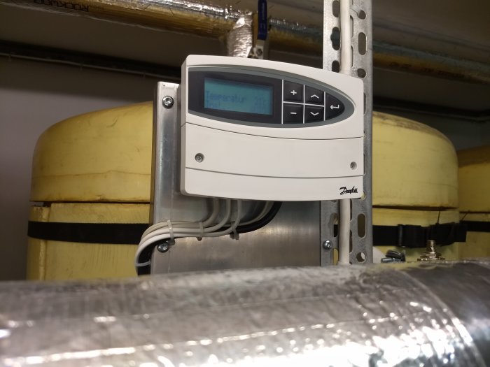 Danfoss ECA61 reglerpanel monterad på väggskena i pannrum, visar temperatur på displayen, med isolerade rör i bakgrunden.