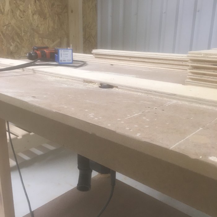 Arbetsbänk med spånsug och verktyg i en verkstad med plywoodskivor i bakgrunden.