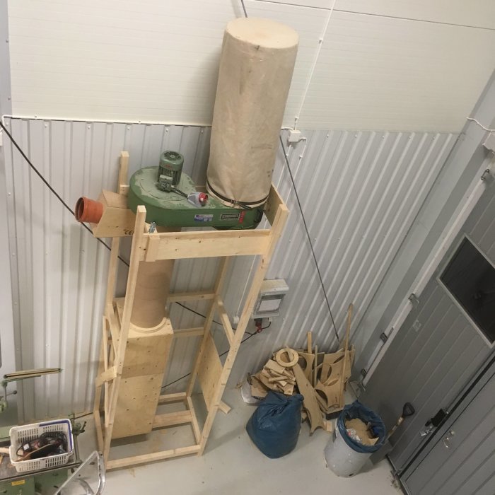 En hemgjord träcyklon med fläkt installerad på ställning i ett verkstadsutrymme, med överskottsmaterial runtomkring.