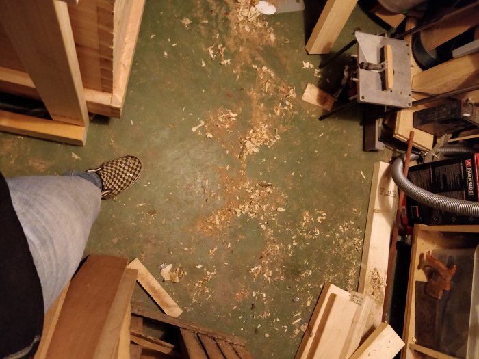 Person står i ett verkstadsrum med träspån på golvet och byggmaterial omkring.