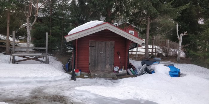 Röd verktygsbod i snölandskap med diverse föremål utanför, dörren stängd.