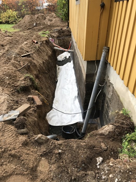 Nybyggd källarvägg med dräneringsarbete, synliga dräneringsrör och plastmatta vid husgrund.