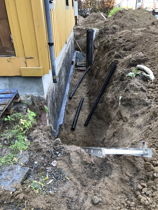 Ny källarvägg med betongputs vid grävd grund nära gult hus, med dräneringsrör och grushög.