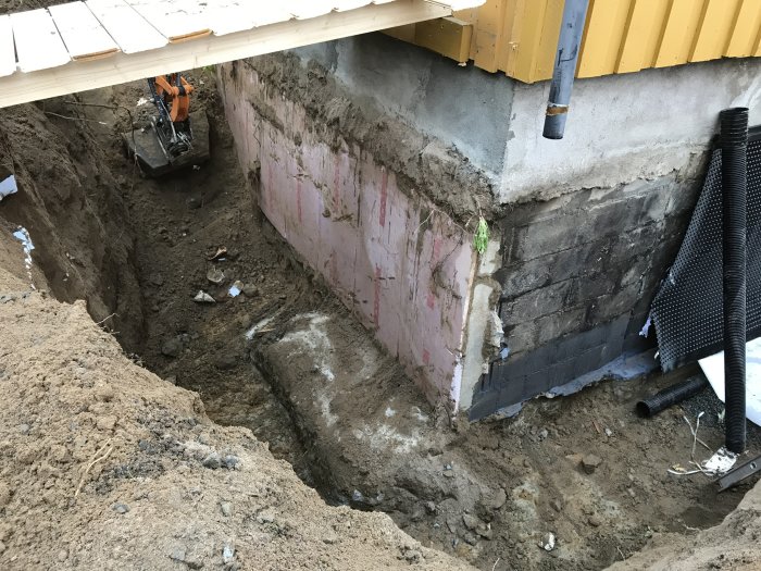 Öppen grund till ett hus med synlig ny och gammal källarvägg, delvis återfylld grävning och dräneringsrör.