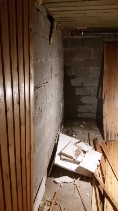 Ett blottat hörn av en källarvägg med stora sprickor och spår av fukt, träpanel och röran på golvet.