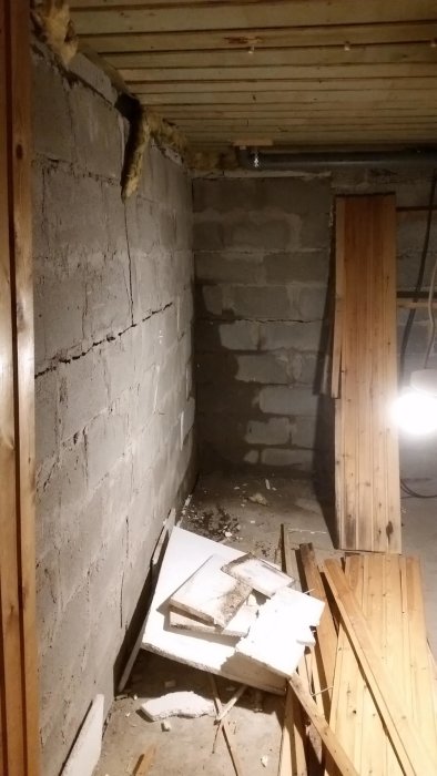 En källarhörna med en stor spricka i väggen och borttagen träpanel samt synliga fuktskador och spillror på golvet.