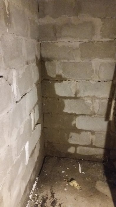Fuktiga betongväggar i ett hörn av en källare med synlig spricka och rester av rivet material på golvet.