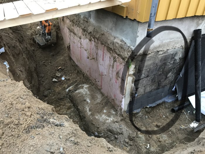 Grävd yta vid husvägg före renovering, isolering synlig på väggen, nedgrävd grön slang och snöhögar.