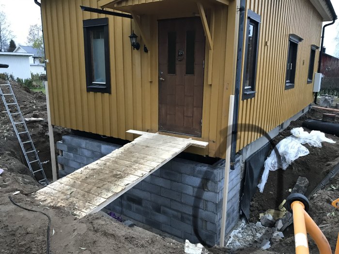 Renoverat hus med ny källargrund av betongblock, trätrappa och gul fasad, byggarbetsplats med material och verktyg synliga.