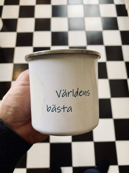 Hand håller en mugg med texten "Världens bästa" framför svartvit rutigt golv.