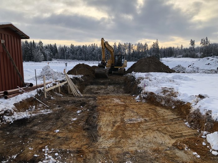 Grävmaskin står vid en utgrävd dunge matjord på en snöig byggarbetsplats med omgivande träd.