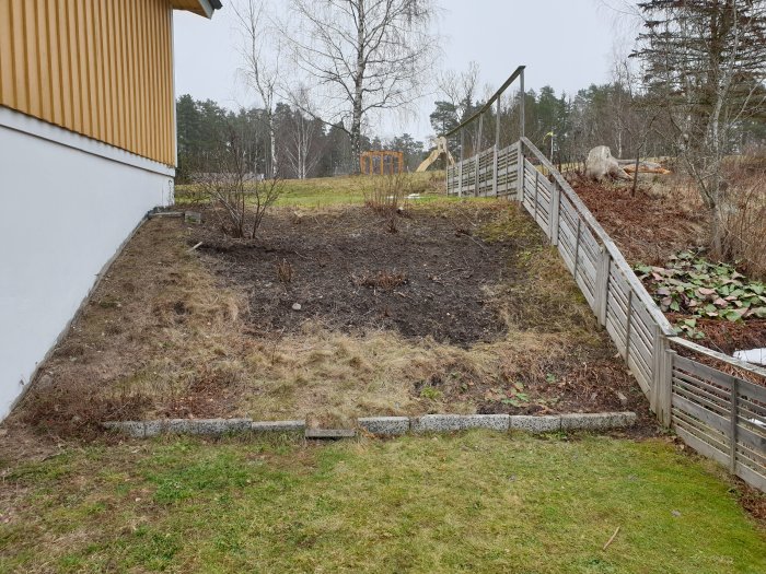 Ojämn slänt med brun gräs och jord intill ett hus, separerad av låg stenmur, med trästaket i bakgrunden.