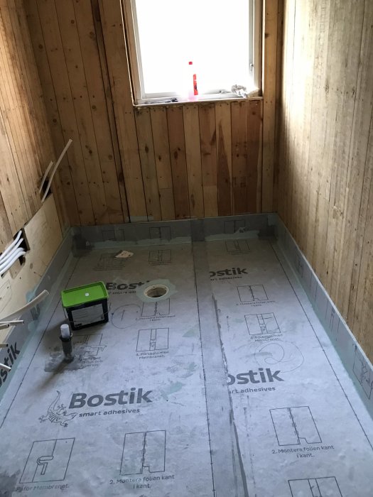 Badrum under renovering med isolerade VVS-rör i golvet, väggar klädda med träpanel och fönster.