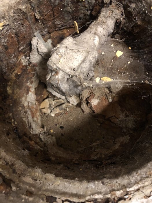 Närbild på en gammal gjutjärnsbrunn med synliga rör och skador, täckt av rost och spindelväv.