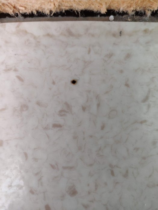 Närbild på en matta med ett litet bränt hål efter ett upphettat gem, intill en vägg med list.
