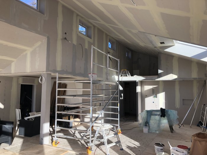 Rum under renovering med gipsskivor på väggar och i tak, spackling synlig, med arbetsställningar och målarutrustning.