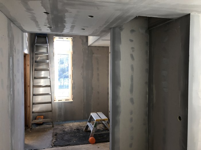 Gipsade väggar och tak med spackelslipning i ett under-renoveringens rum med stege och plywoodvägg.