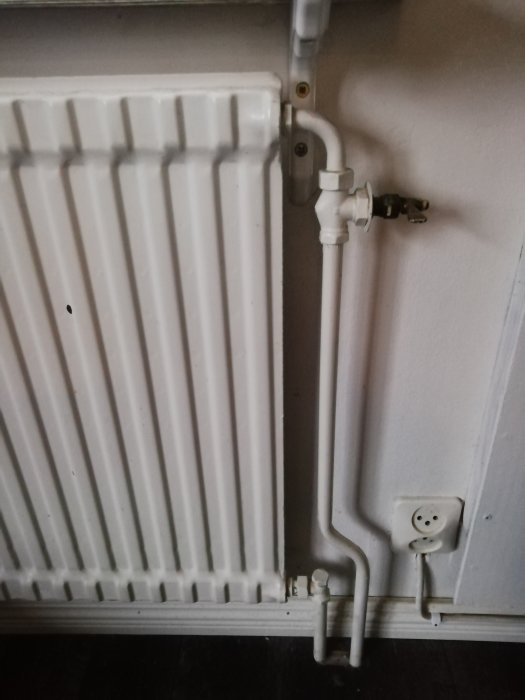Radiator med reglerventil och anslutningsrör i ett rum, bredvid ett eluttag.