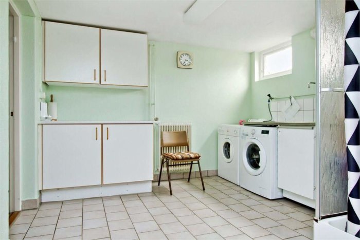Tvättstuga med vita skåp, tvättmaskin, torktumlare och beige klinkergolv före renovering.