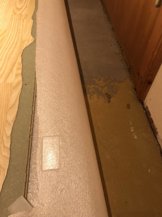 Upprivet laminatgolv som visar underliggande spånskivor med rester av gulgrönt pulveriserat gammalt lim vid kant nära dörr.